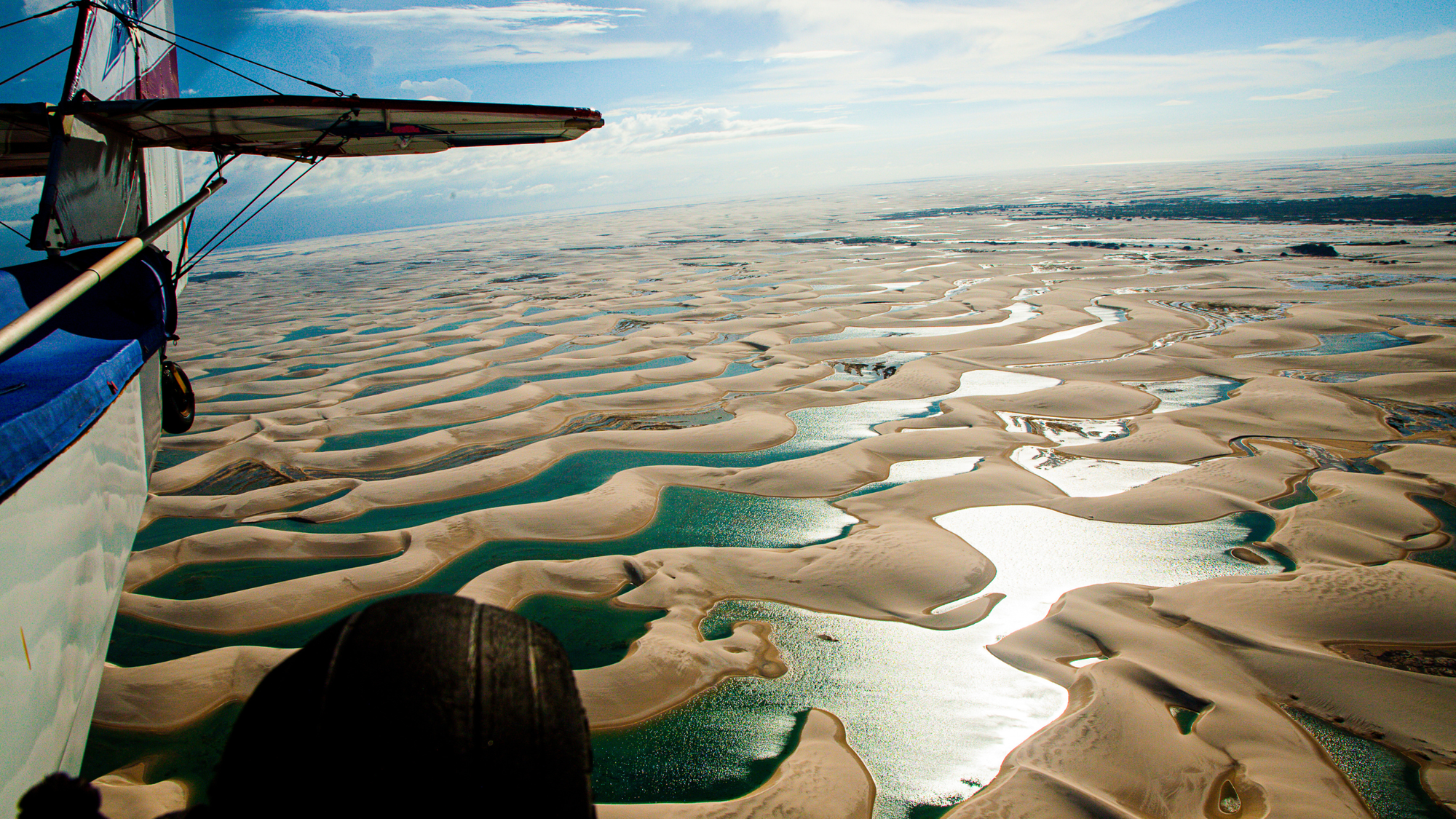 Aerial view from a small airplane flying over Lençóis Maranhenses National Park in Brazil, showing endless white sand dunes and blue lagoons under a partly cloudy sky.