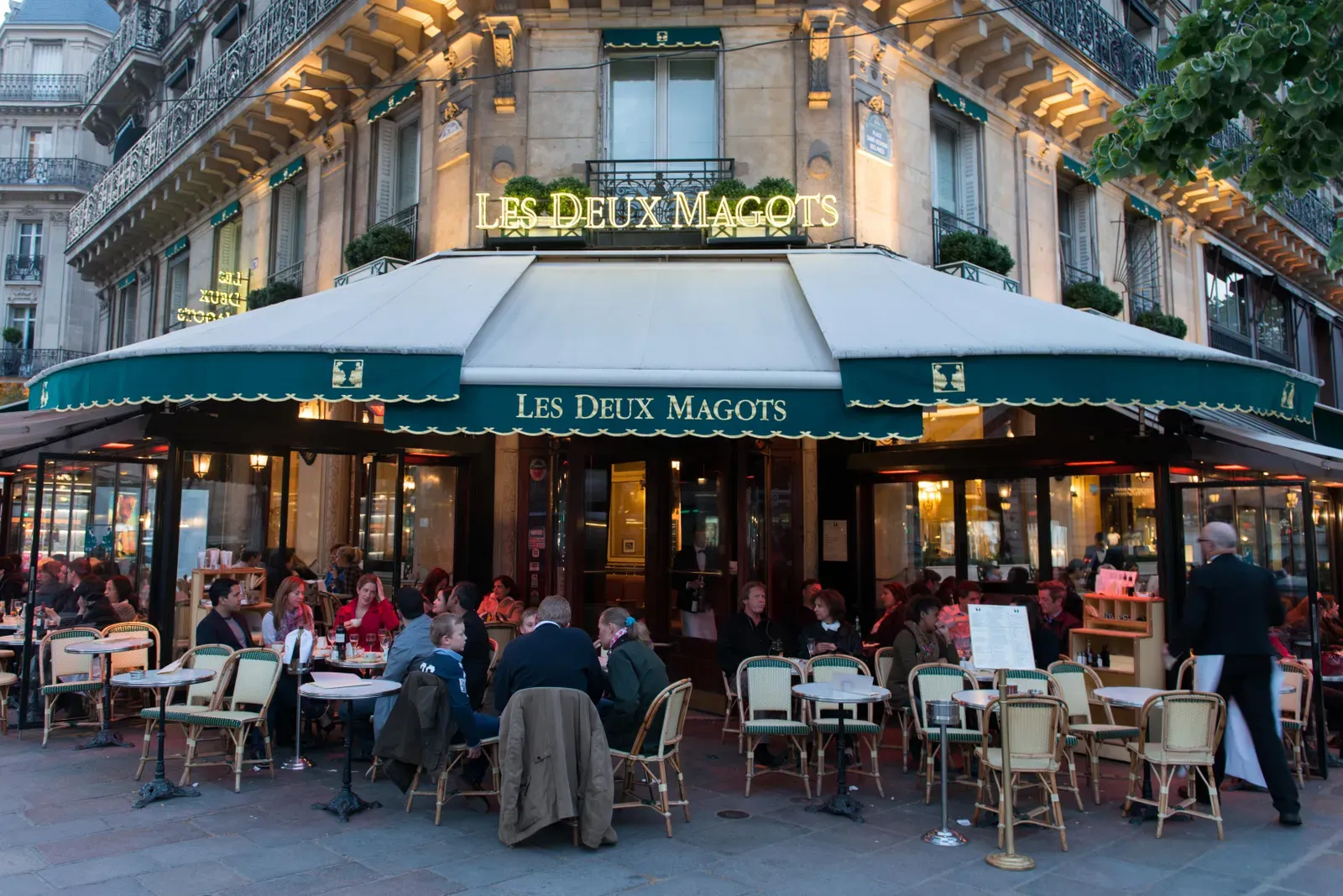 People dining at the iconic Les Deux Magots café in Paris, seated at outdoor tables under a green and white awning on a lively evening.