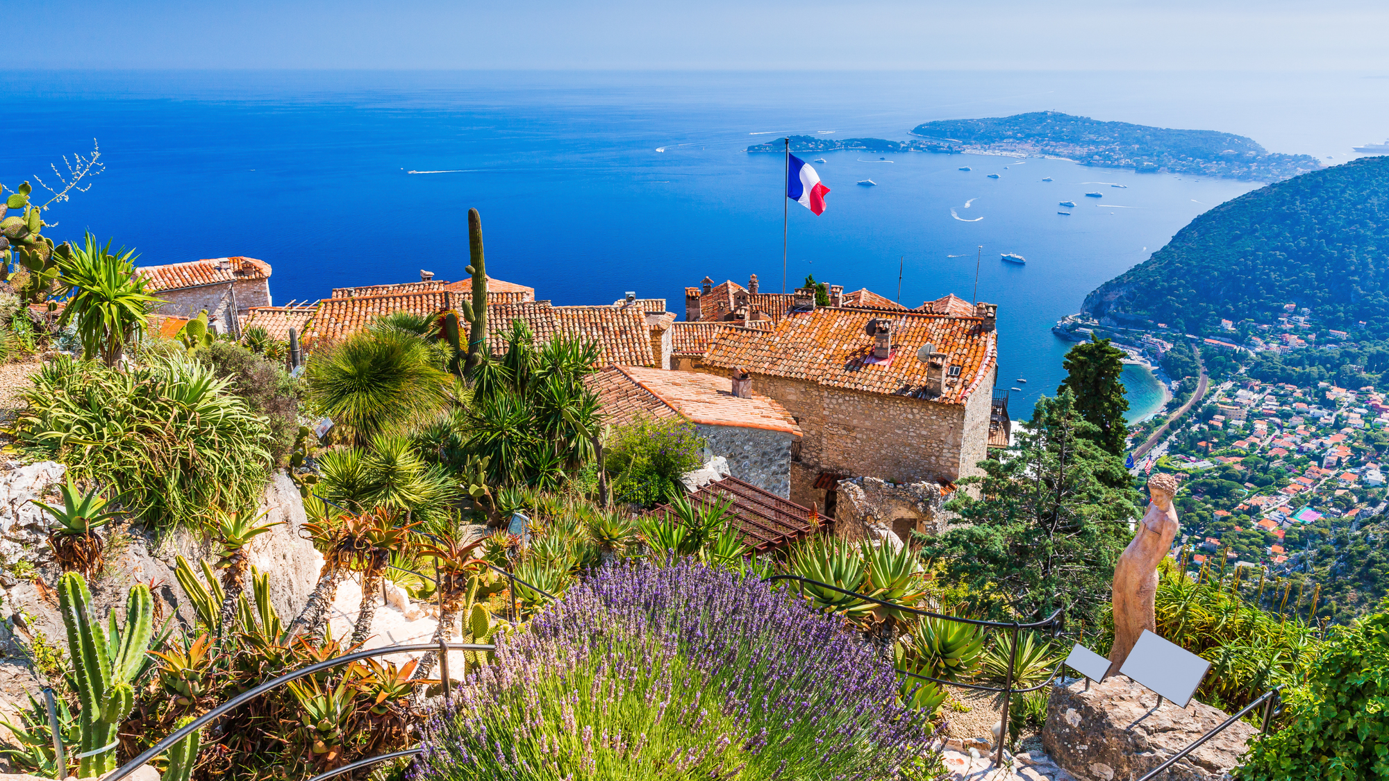 Hilltop view of Èze on the French Riviera with terracotta rooftops, coastal plants, a French flag, and the blue Mediterranean Sea dotted with boats below.