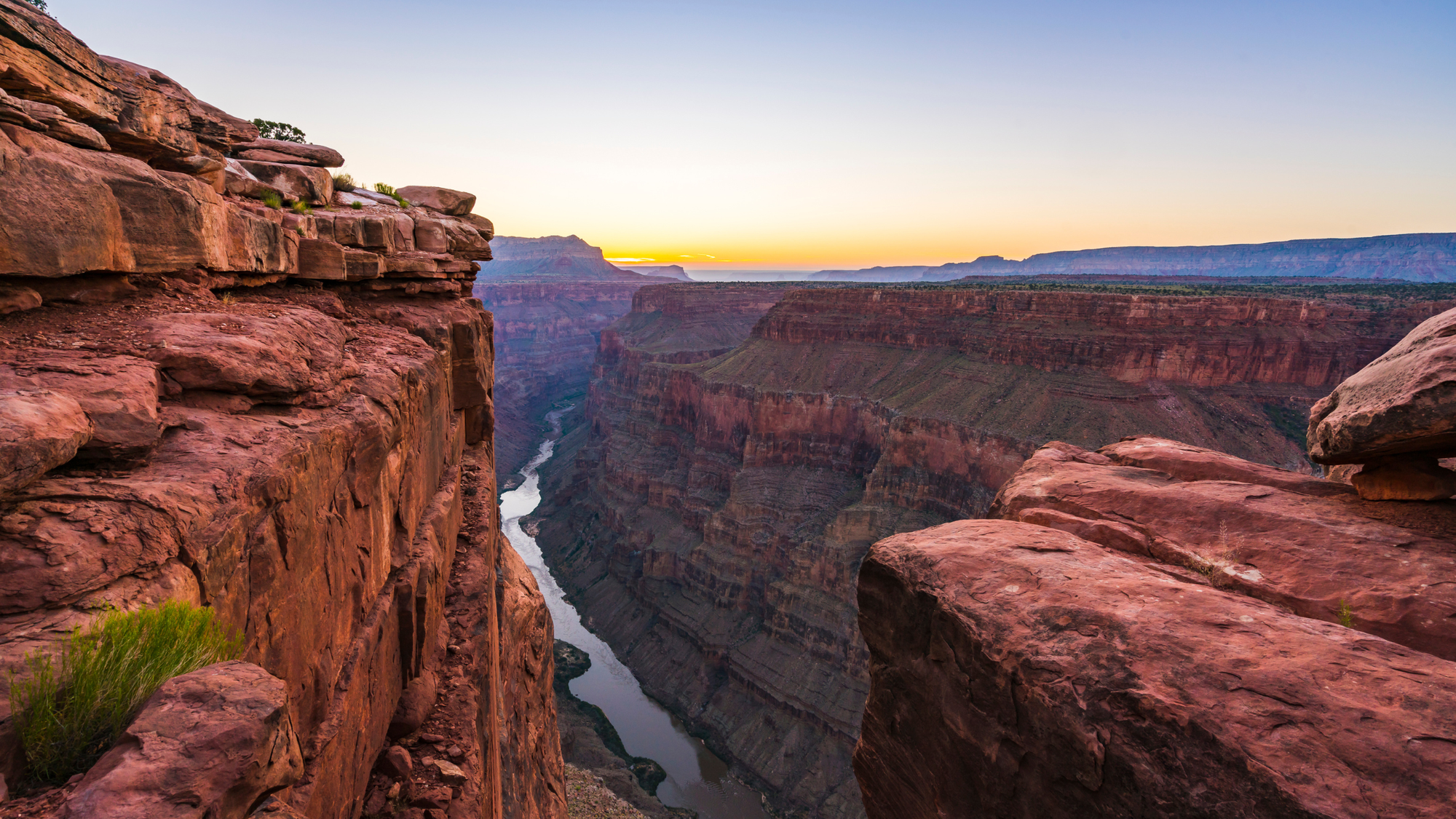 Sunset view from the edge of a red rock cliff overlooking the Colorado River winding through the deep, layered canyons of Grand Canyon National Park, Arizona.