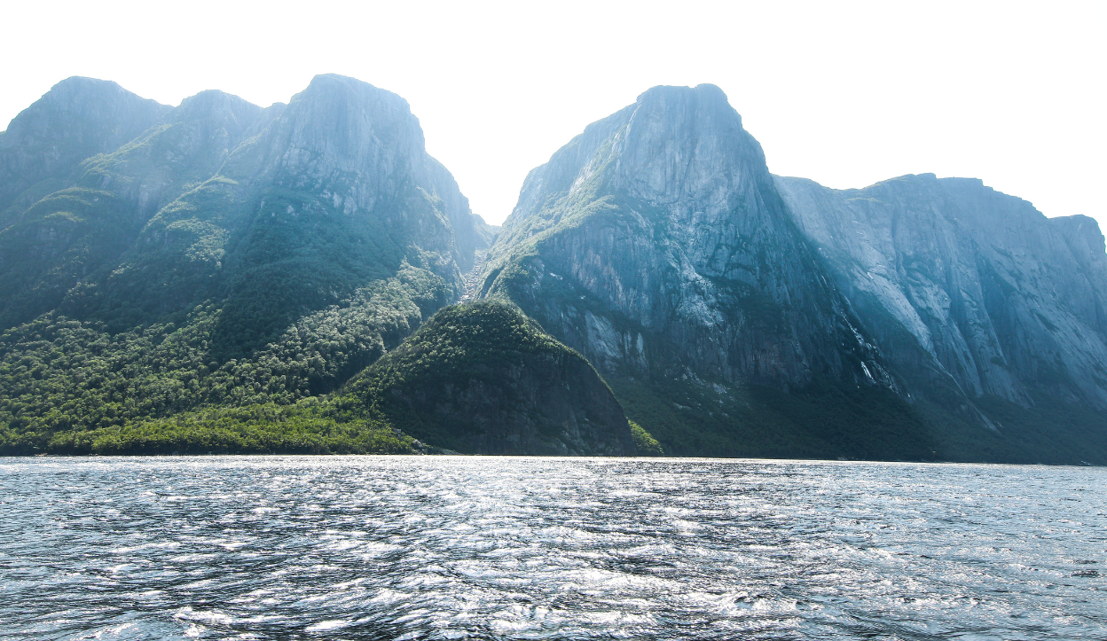 Steep, forested mountains rise above a glistening body of water under bright sunlight in Gros Morne National Park, Newfoundland and Labrador, Canada.