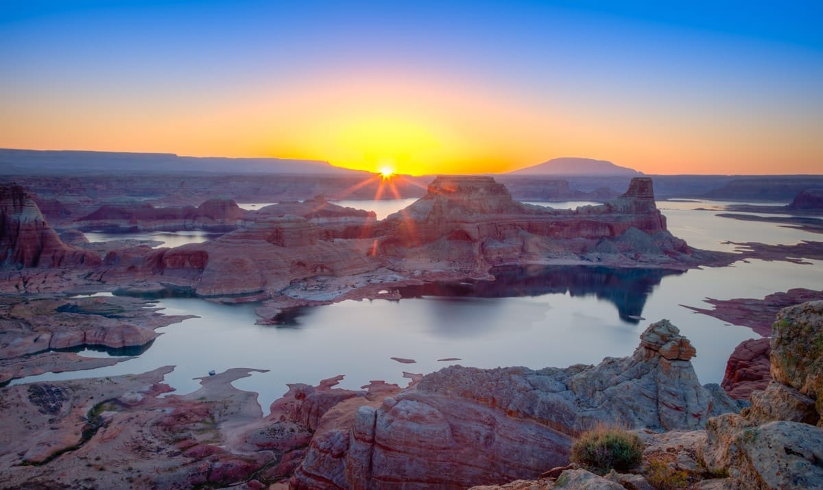 A glowing sunset over Lake Powell lights up the red rock formations and winding water channels in Gunsight Canyon.