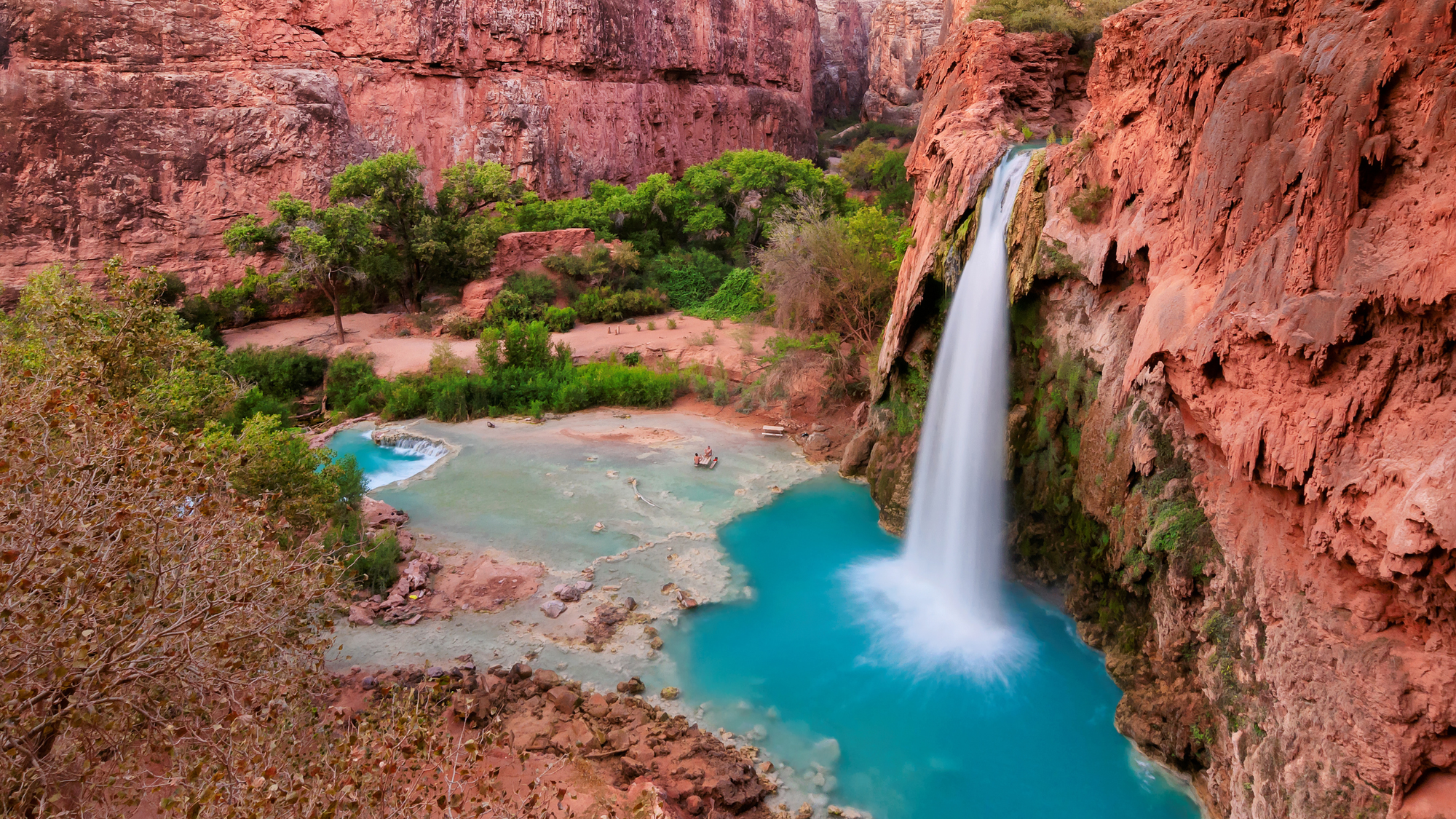 Scenic view of Havasu Falls cascading over red rock cliffs into a turquoise blue pool surrounded by lush green trees and vegetation in the Grand Canyon, Arizona.