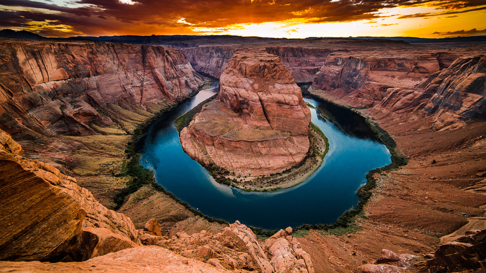 Dramatic view of Horseshoe Bend in Arizona at sunset, showing the Colorado River curving around a large sandstone rock formation, with steep red canyon walls and a vibrant sky in the background.