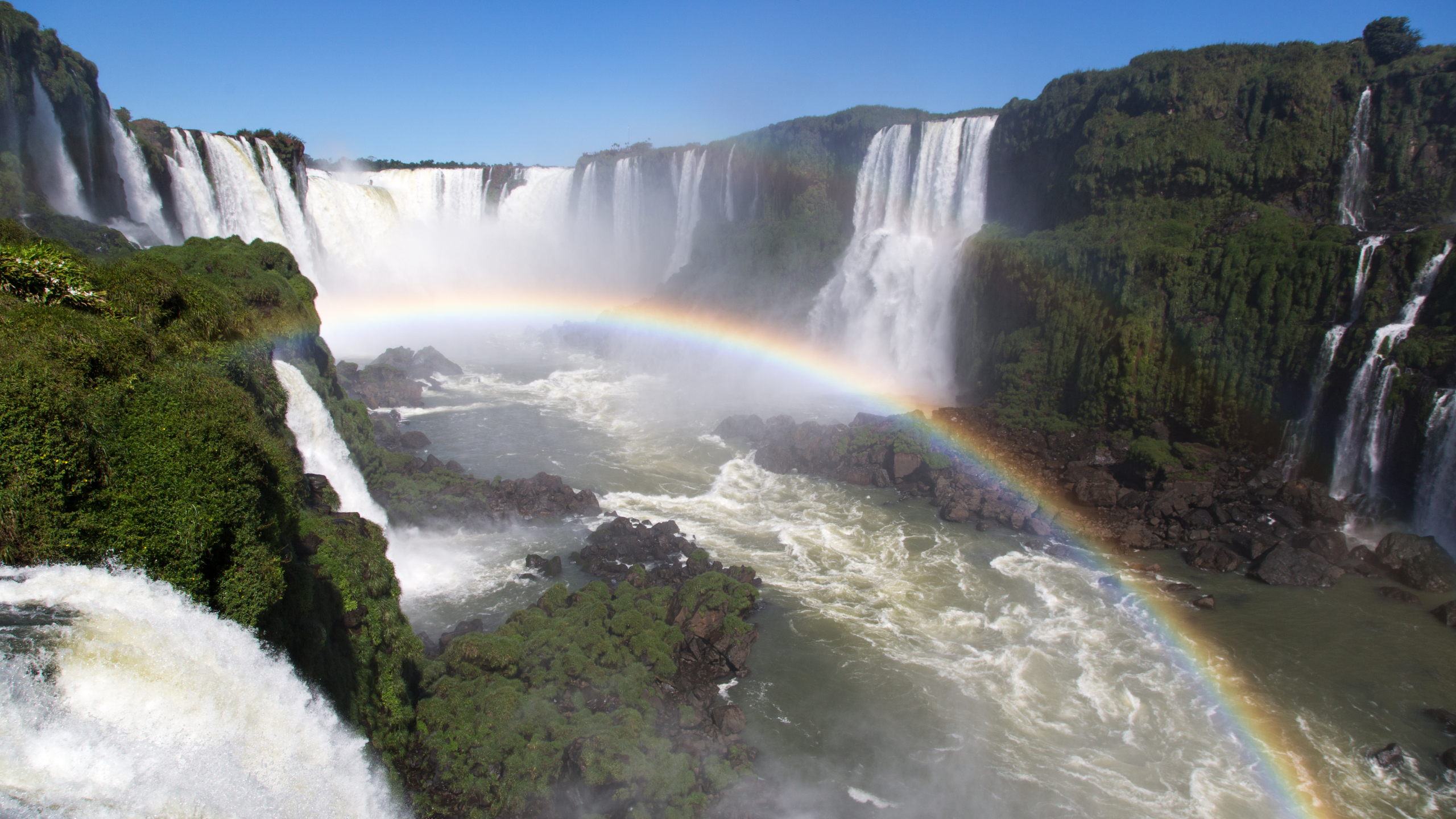 Powerful waterfalls cascade over lush, green cliffs in Iguaçu National Park in Brazil, with mist rising and a vibrant rainbow arching over the rushing river.