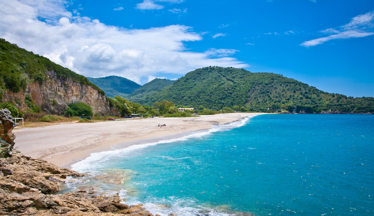 Wide sandy beach at Karavostasi in Epirus, Greece, bordered by turquoise waters and lush green hills under a partly cloudy blue sky.