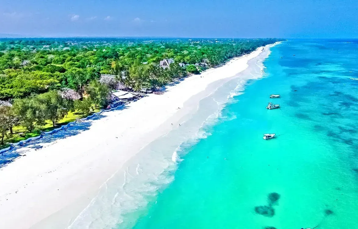 Aerial shot of Kilifi Beach, Kenya, displaying a pristine white sand shoreline bordered by dense tropical vegetation and calm, turquoise-blue ocean waters with anchored boats.