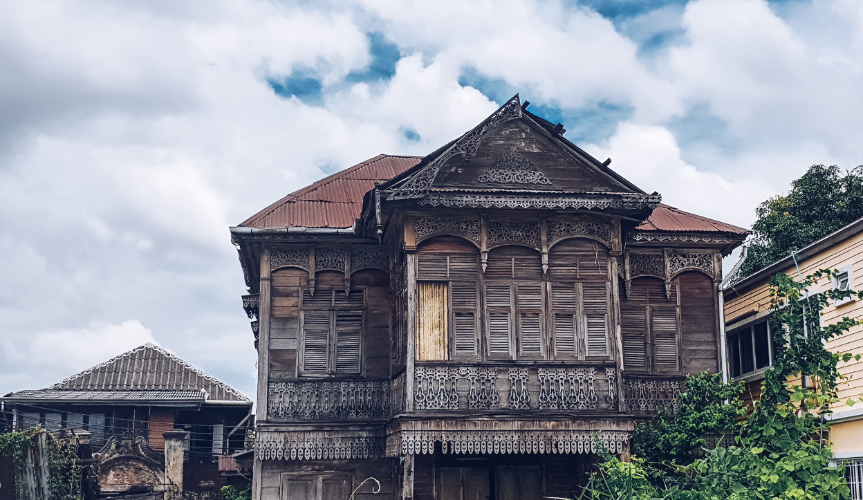 Old wooden Thai-Portuguese style house in the historic Kudi Chin community in Bangkok, with ornate wooden carvings, shuttered windows, and a rusted metal roof, surrounded by nearby homes and greenery.