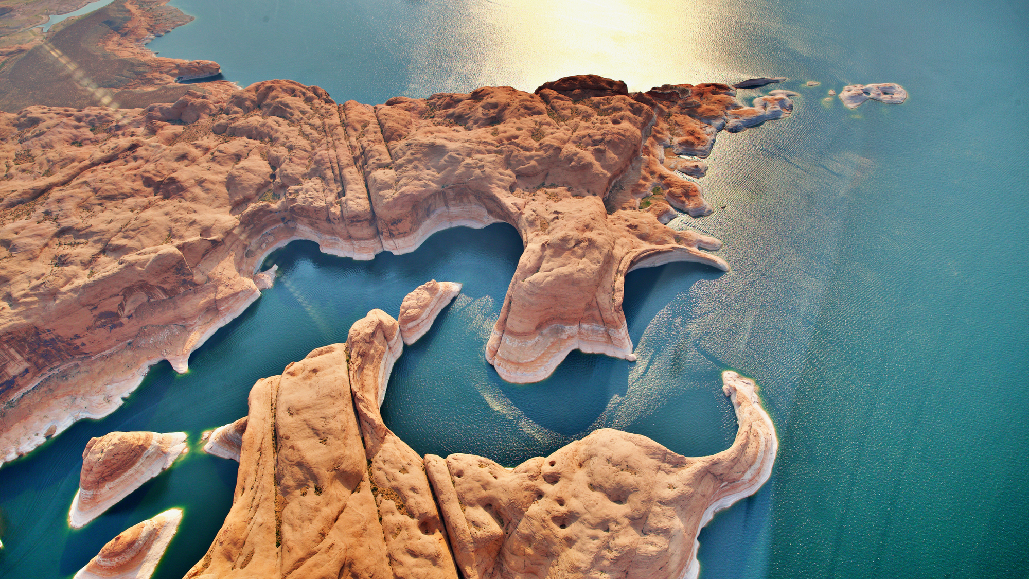 Aerial view of the red sandstone formations and winding shoreline of Lake Powell in Arizona, with deep blue water and sunlight reflecting off the lake’s surface.