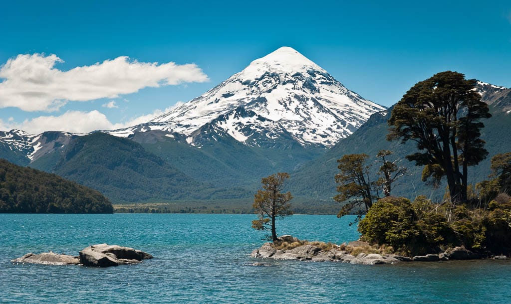 Snow-capped Lanín Volcano rises behind a pristine lake and wooded islands in Lanín National Park, Argentina, under a vibrant blue sky with scattered clouds.