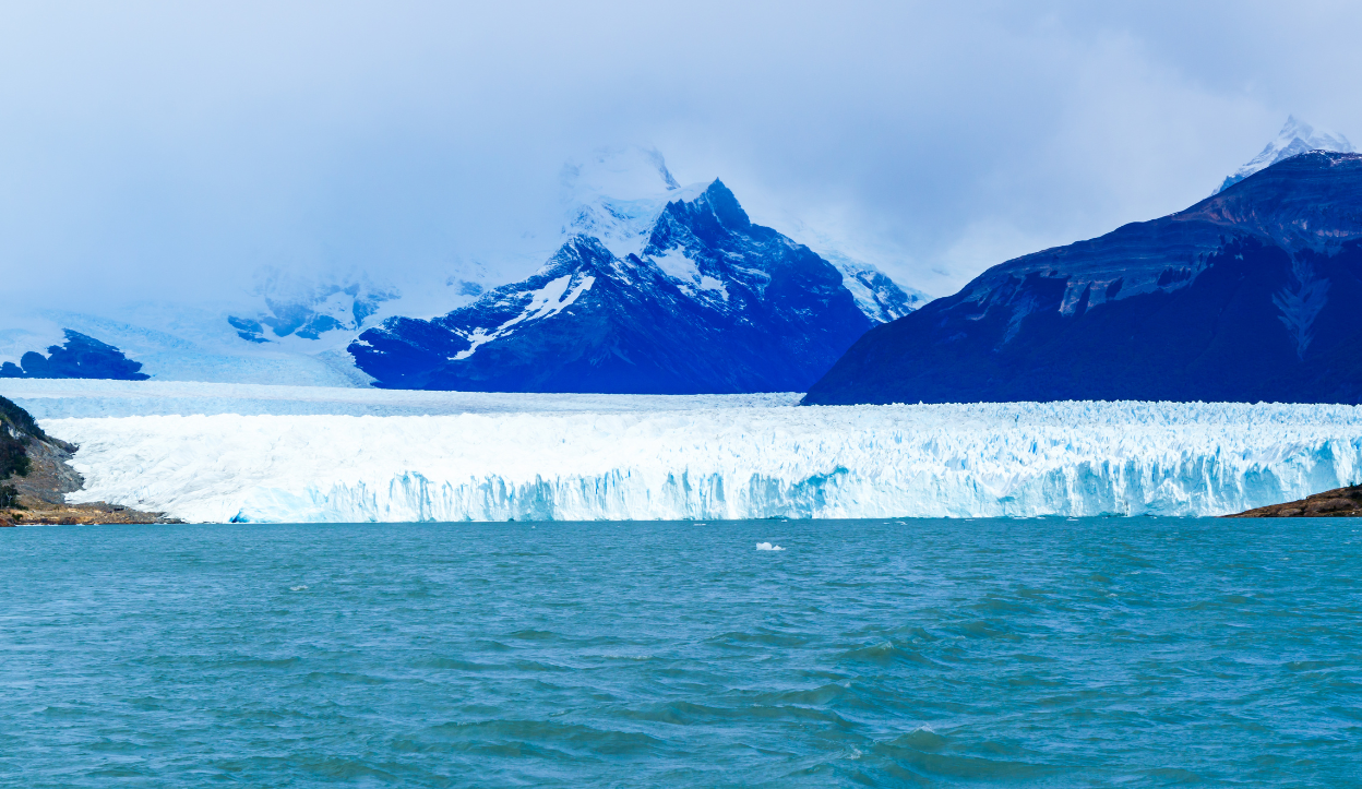 Massive wall of blue ice from a glacier meets turquoise water in Los Glaciares National Park, Argentina, with snow-covered peaks and misty clouds in the background.