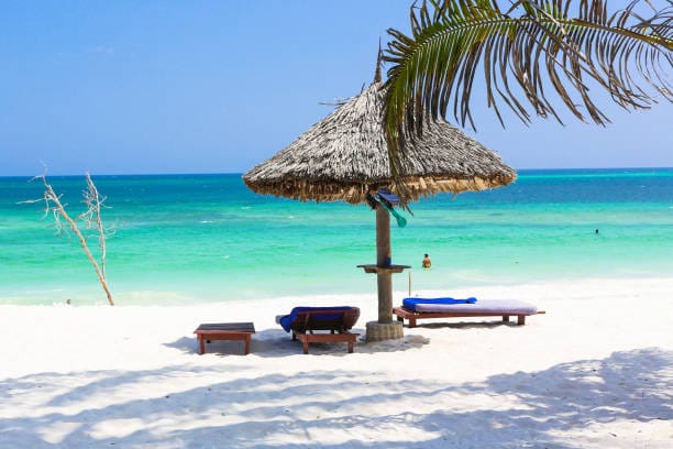 Tranquil beachfront scene in Malindi, Kenya, featuring a thatched sunshade, wooden lounge chairs on white sand, and turquoise sea waves under a sunny blue sky.