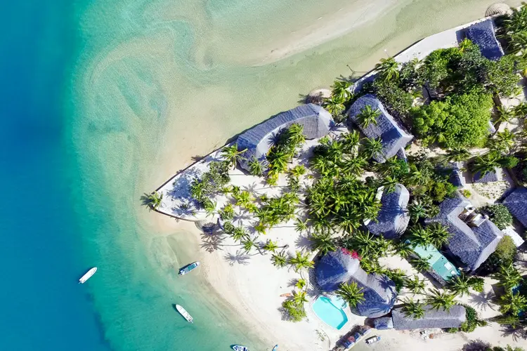 Overhead view of Manda Bay on Lamu Island, Kenya, with thatched villas, coconut palms, white sand beach, and boats floating in the clear blue-green waters.