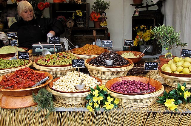 Colorful market stall at Montmartre Grape Harvest Festival in Paris, displaying baskets of olives, sun-dried tomatoes, garlic, and other local produce with handwritten signs.