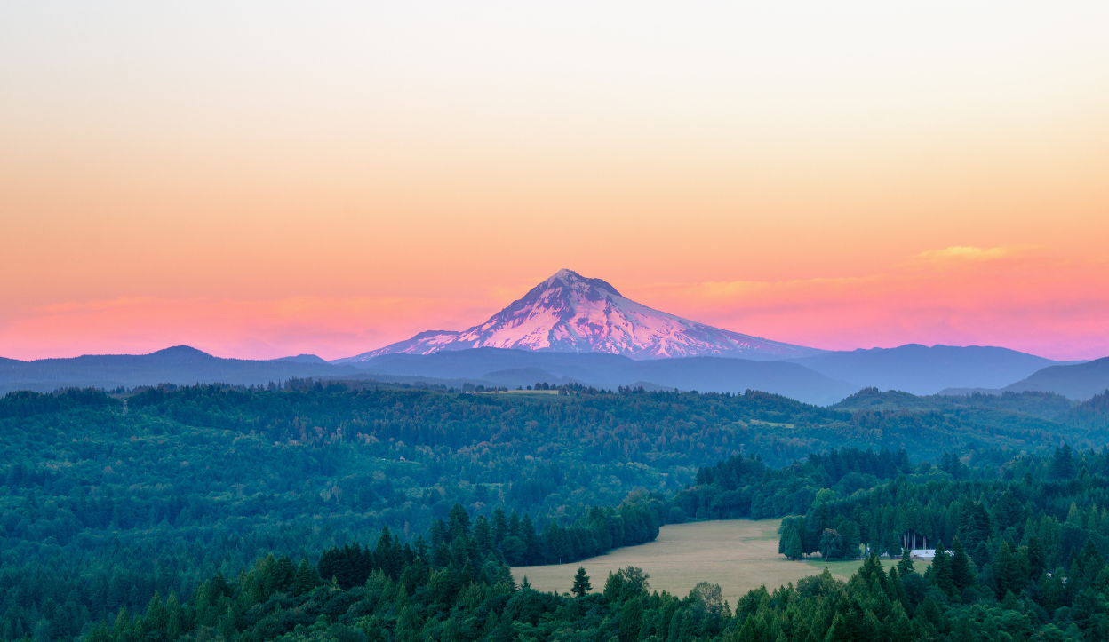 Majestic view of Mount Hood with snow-covered slopes at sunrise, framed by layers of forest and open meadows in Mount Hood National Forest, Oregon, under a colorful pink and orange sky.