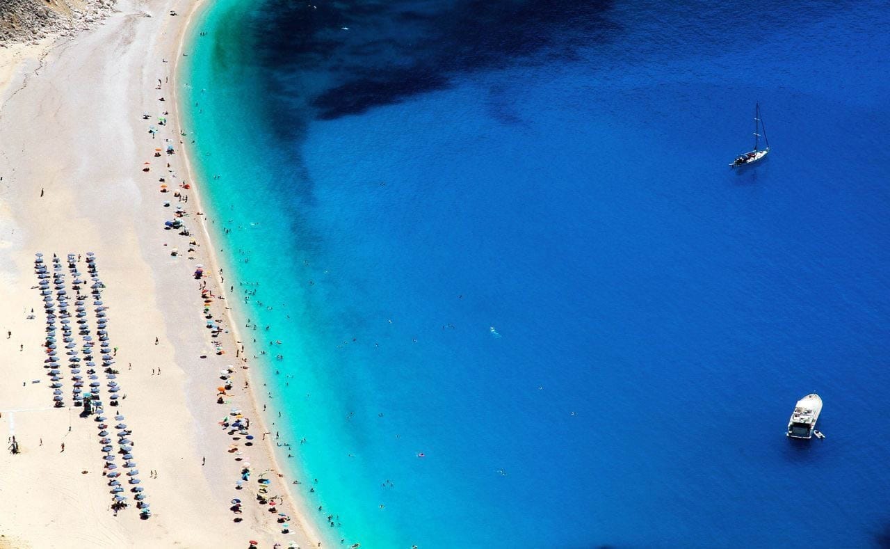 Aerial view of Myrtos Beach on Kefalonia Island, Greece, featuring white pebbled shores, deep blue water, and colorful umbrellas lined along the beach.