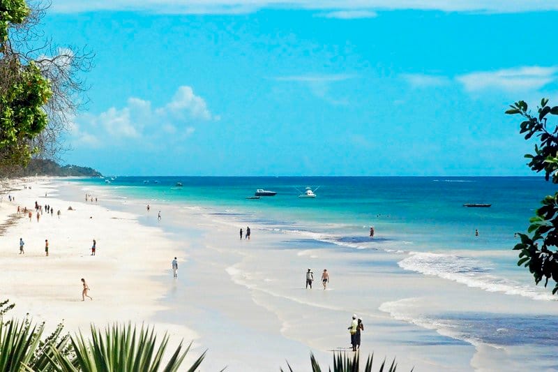Nyali Beach in Mombasa, Kenya, with tourists walking along the wide stretch of white sand, bordered by lush vegetation and the blue waters of the Indian Ocean dotted with boats.
