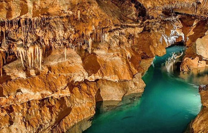 Underground stream flowing through Onyx Cave in Arkansas, with illuminated turquoise water and orange rock formations with stalactites.