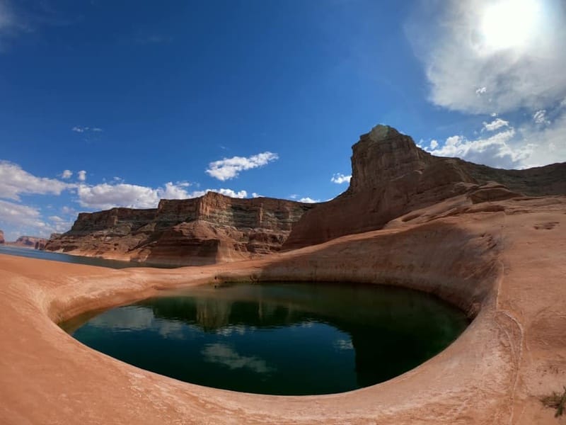 Circular water-filled rock formation known as the Toilet Bowl sits near Padre Bay at Lake Powell under a partly cloudy sky.