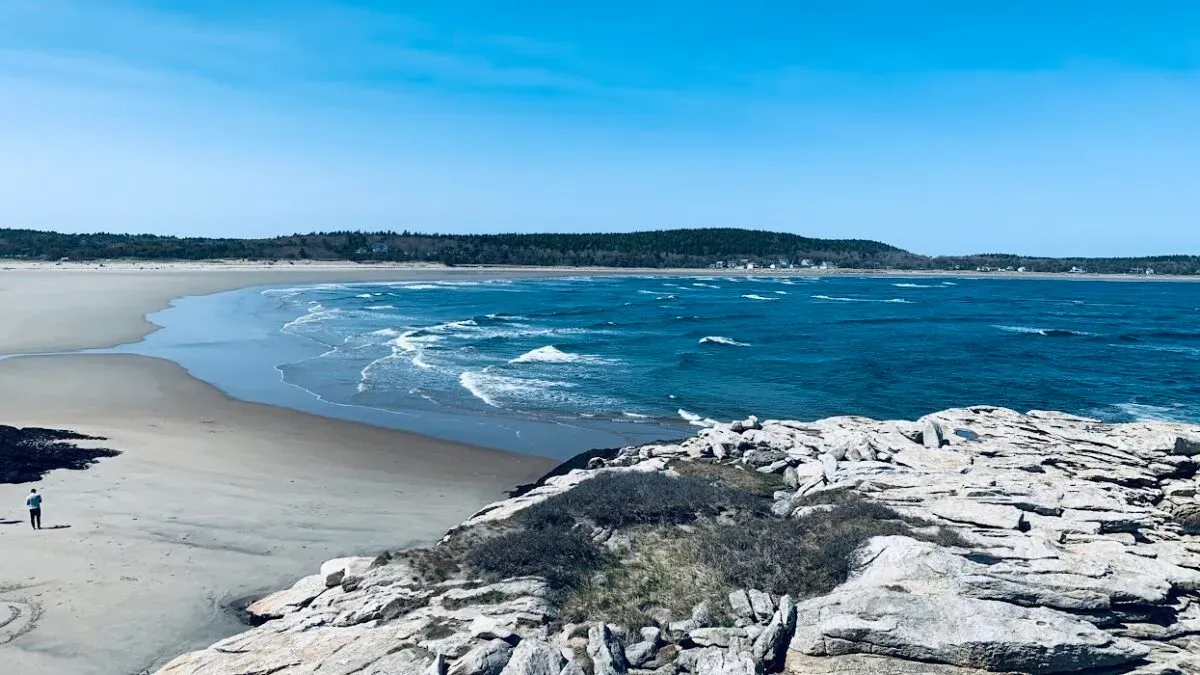 Aerial view of Popham Beach in Phippsburg, Maine featuring rugged white rock formations meeting the ocean, with a wide sandy beach and forested marshland inland.