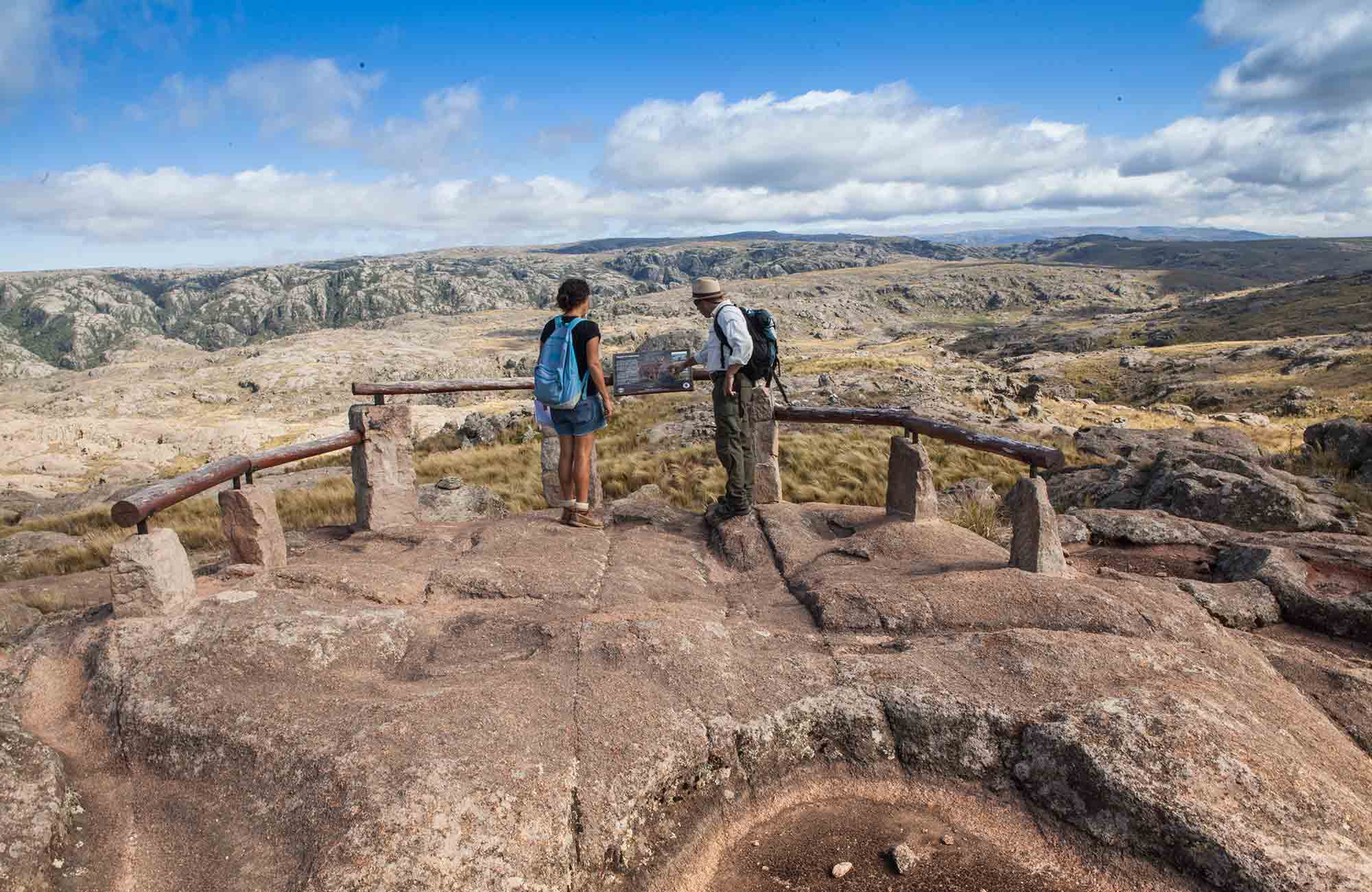Two hikers stand on a rocky viewpoint overlooking the vast rugged canyons and grasslands of Quebrada del Condorito National Park in Argentina under a partly cloudy sky.