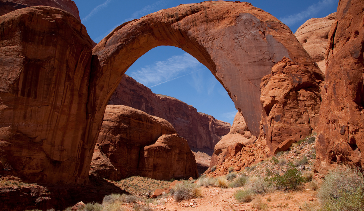 Massive natural stone arch of Rainbow Bridge rises above desert terrain under a clear blue sky at Lake Powell.