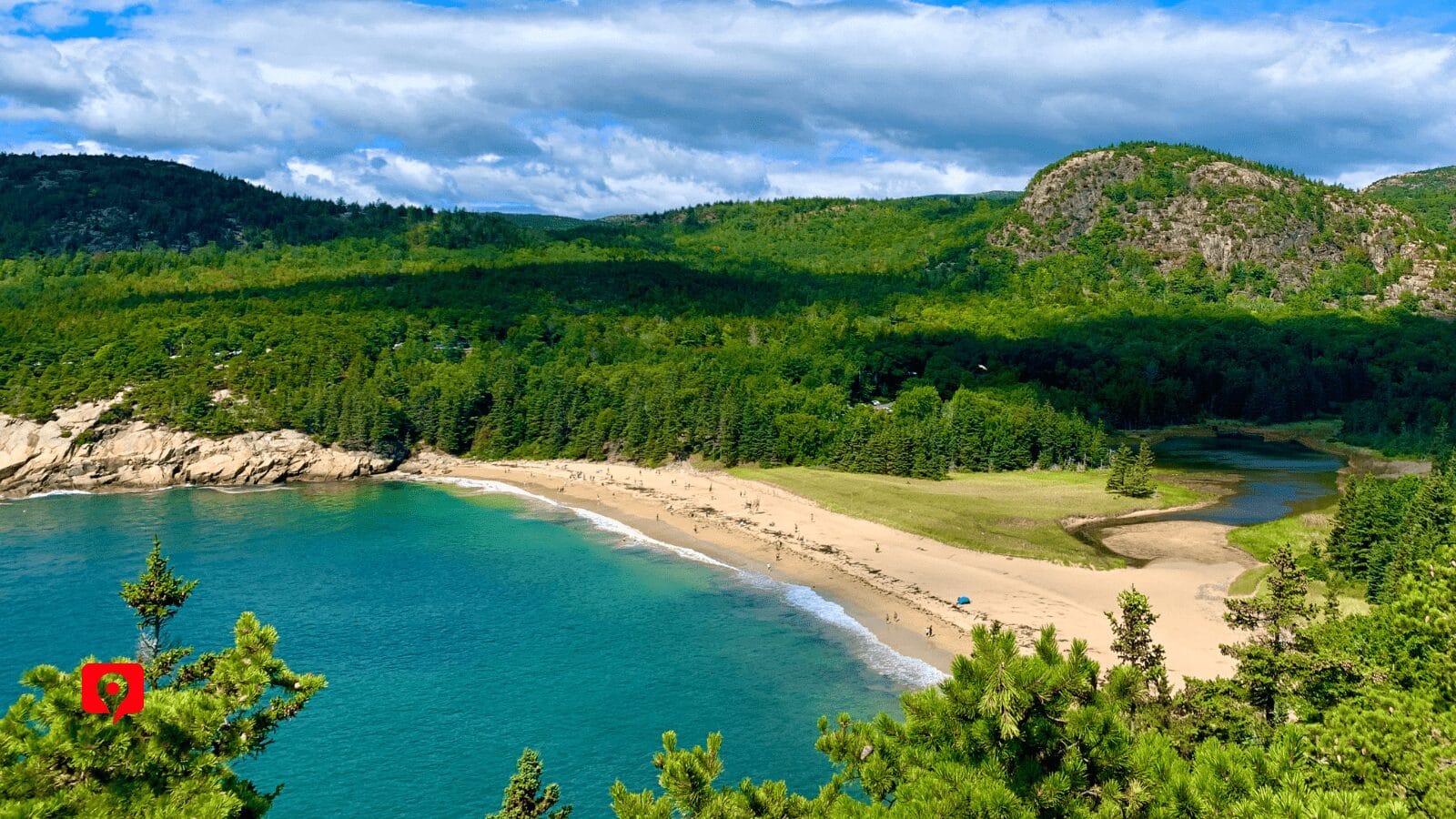 Scenic coastal view of Sand Beach in Acadia National Park, Maine, surrounded by pine-covered hills and mountains, with turquoise water gently meeting the crescent-shaped sandy shore.