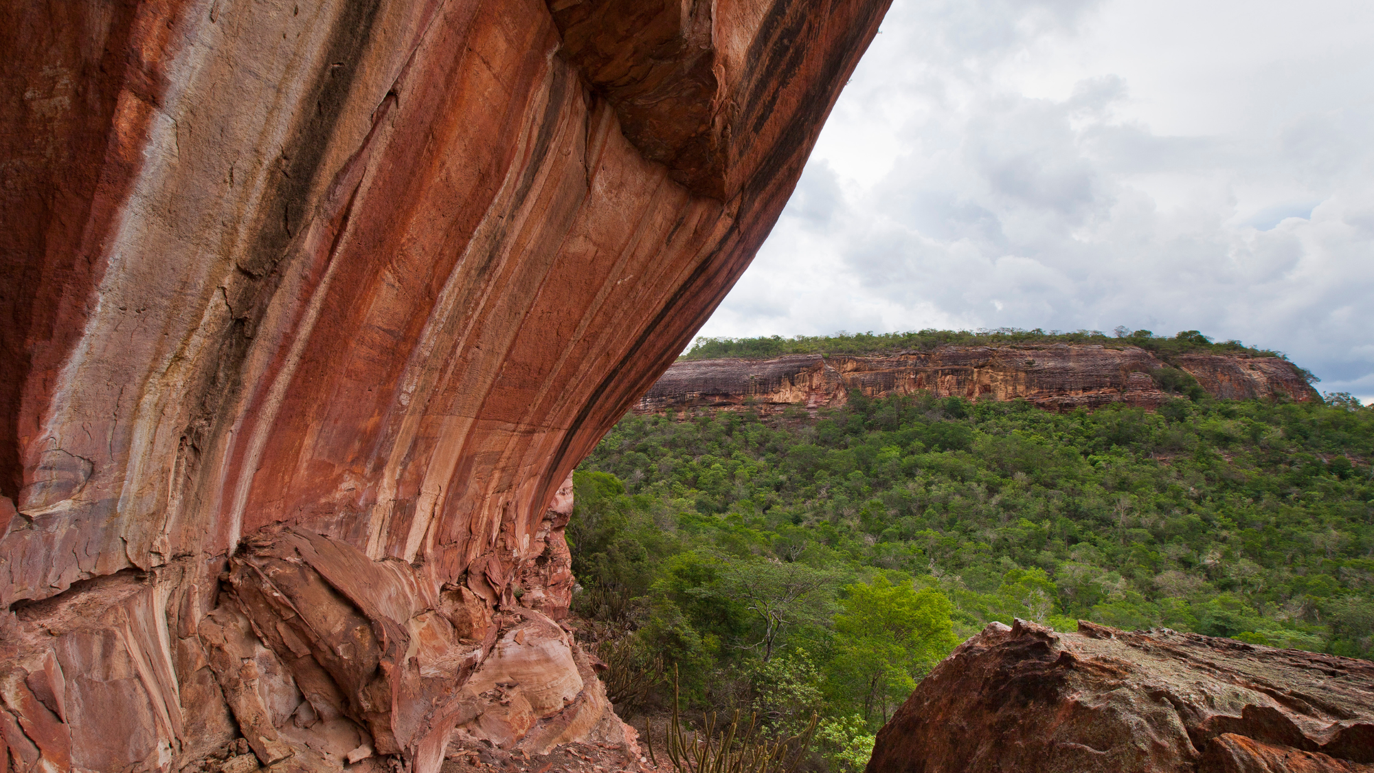 Large red and orange striped rock cliff overlooking green forested hills and distant rocky plateaus under a cloudy sky at Serra da Bocaina National Park in Brazil.