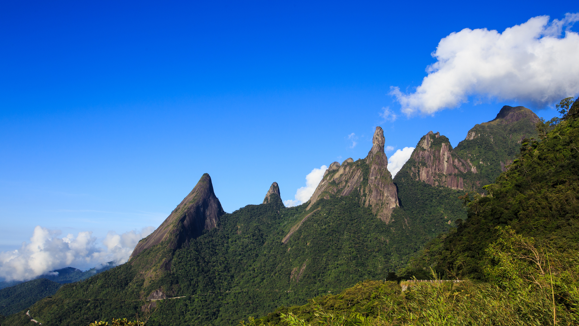 Distinctive jagged peaks covered in green forest rise under a bright blue sky with scattered clouds at Serra dos Órgãos National Park in Brazil.