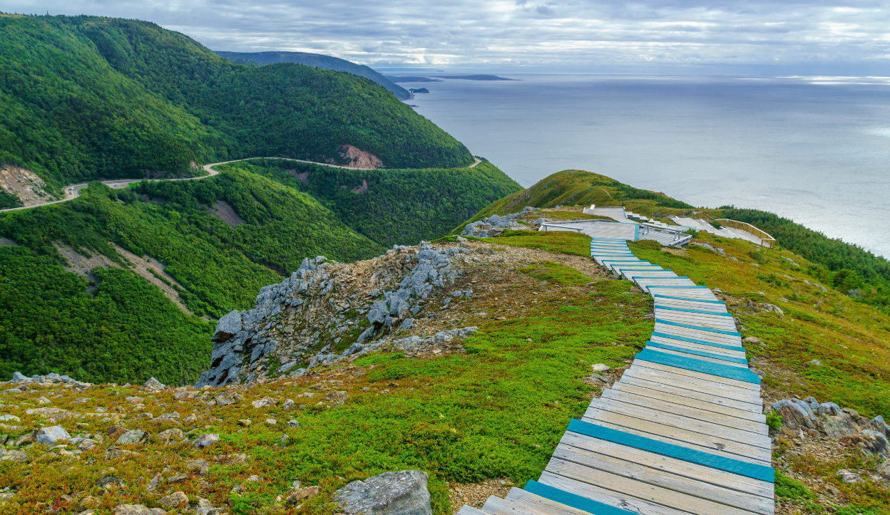 Wooden boardwalk winding along a cliffside overlooking the ocean and rolling green hills on the Skyline Trail in Cape Breton Highlands National Park, Canada.
