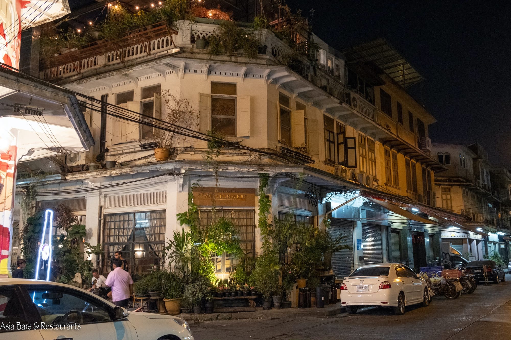 Night view of Soi Nana in Bangkok’s Chinatown featuring a colonial-style shophouse with open windows, potted plants, rooftop string lights, and people socializing near a cozy cafe.