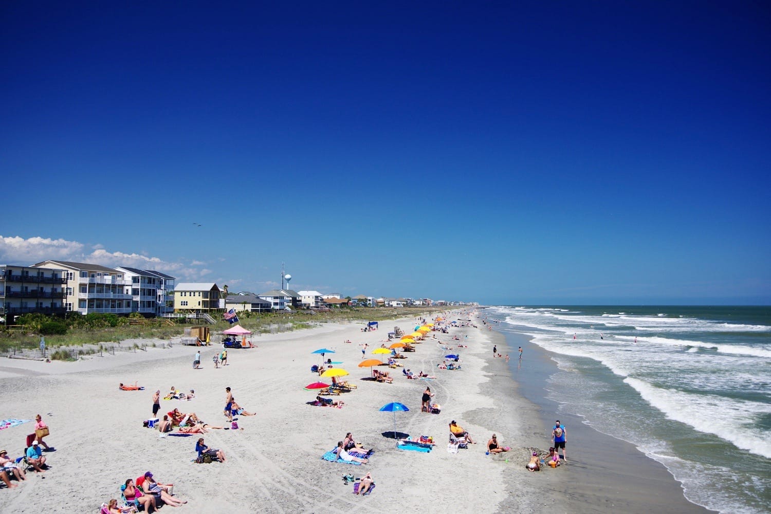 Aerial photo of Sullivan’s Island Beach in South Carolina, lined with upscale homes, natural dunes, and a sandy beach meeting calm ocean waves on a clear day.