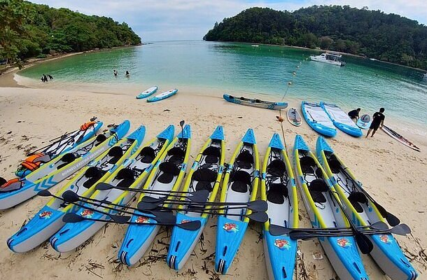 Paddleboards lined up on the sandy shore of Tanjung Aru Beach in Malaysia, with calm turquoise water and green forested hills in the background.