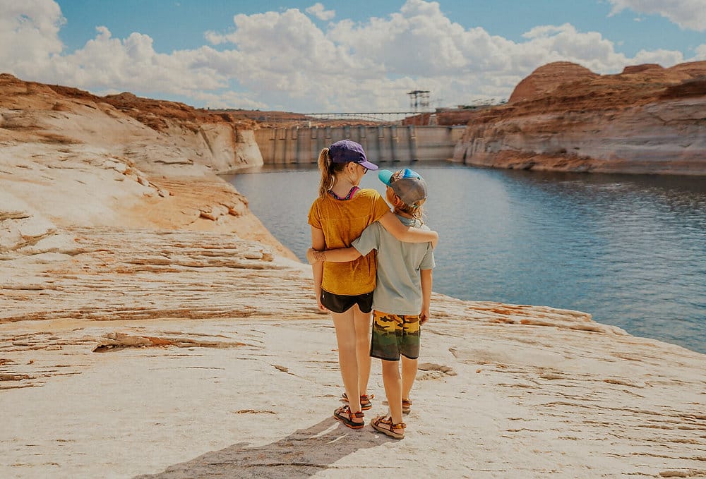 Two children stand arm-in-arm on rocky terrain overlooking Lake Powell near The Chains, with the Glen Canyon Dam in the distance.