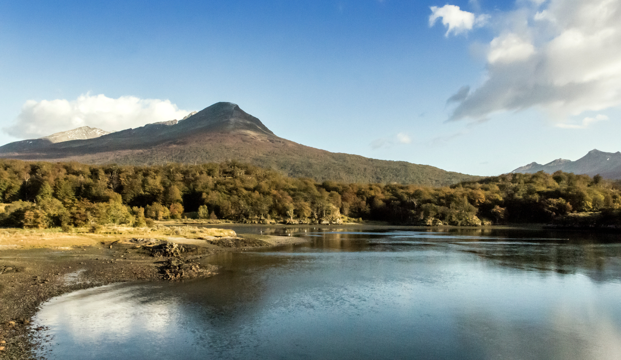 Calm river or lake reflecting surrounding autumnal forest and mountains in Tierra del Fuego National Park, Argentina, with a snow-capped peak in the distance.