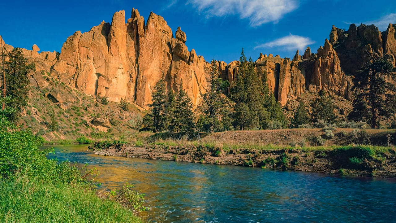 Vibrant landscape of Tumalo State Park in Oregon featuring a bright blue river bordered by grassy banks and pine trees, with striking orange rock cliffs rising under a clear blue sky.