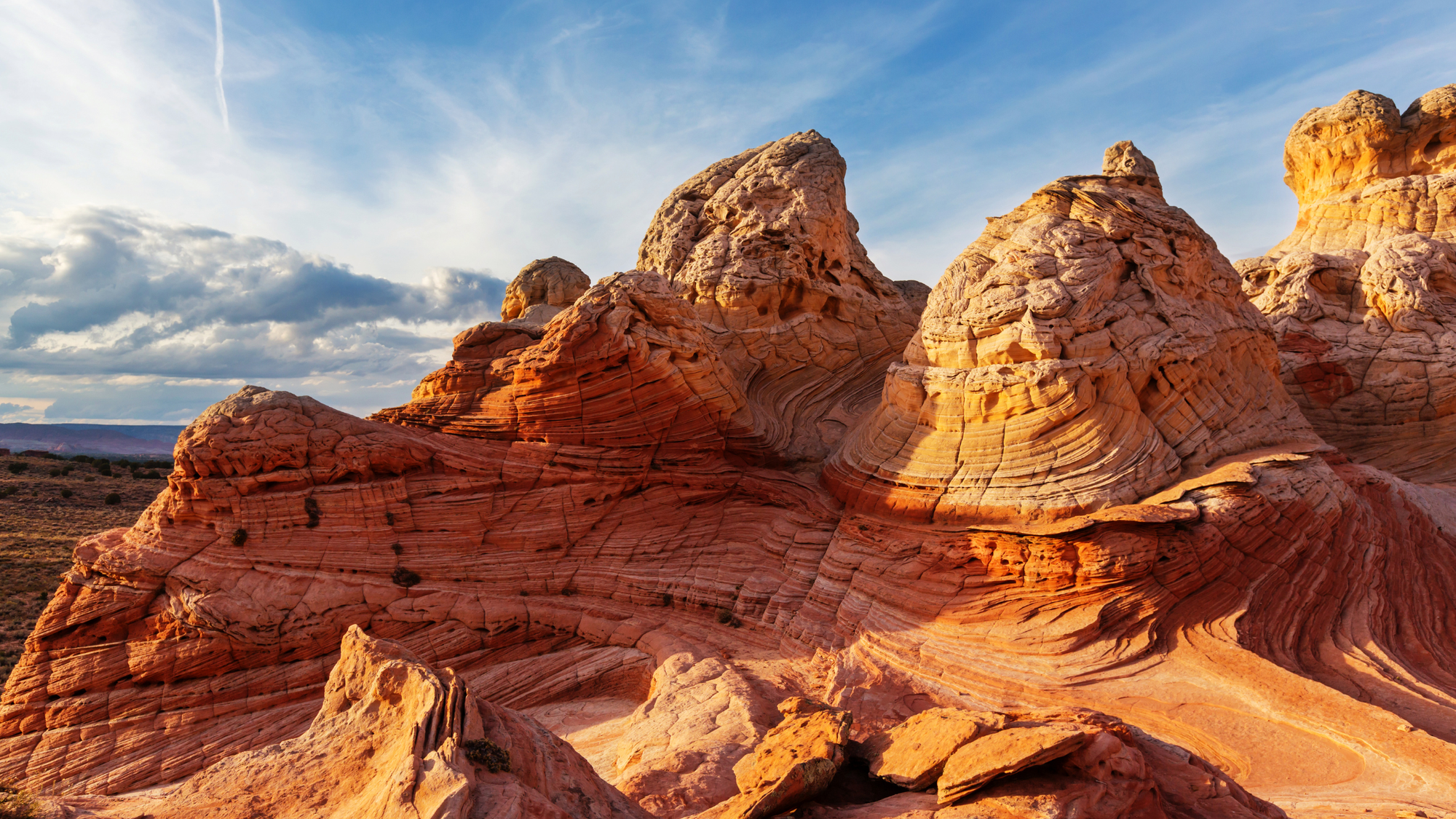 Sunlit sandstone formations with dramatic red and orange swirling layers at Vermilion Cliffs National Monument in Arizona, under a blue sky with scattered clouds.