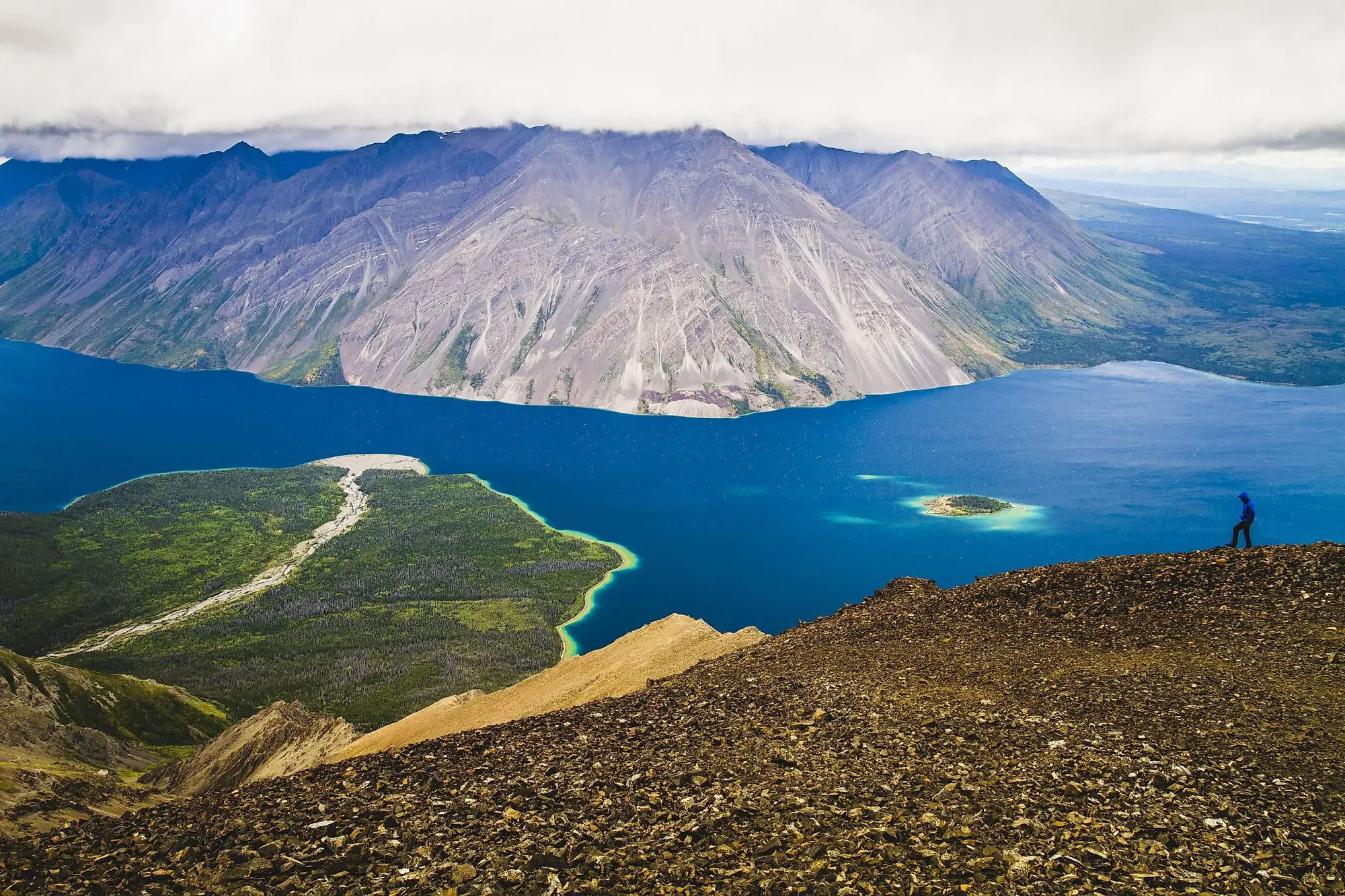 A hiker stands on a rocky slope overlooking a vibrant blue lake and towering mountain range in Kluane National Park, Canada.
