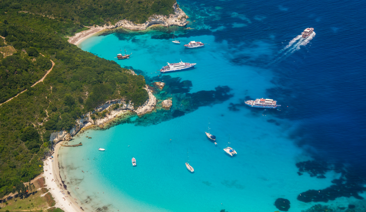 Aerial view of Voutoumi Beach on Antipaxos Island, Greece, showing turquoise waters, anchored boats, and lush green cliffs surrounding the secluded beach.