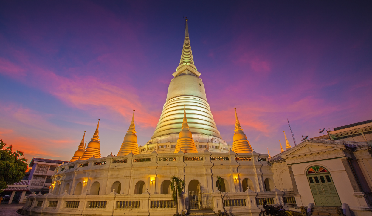 Twilight view of Wat Prayoon's main white chedi glowing under colorful sunset skies in Bangkok, Thailand, surrounded by smaller golden spires and colonial-style temple buildings.