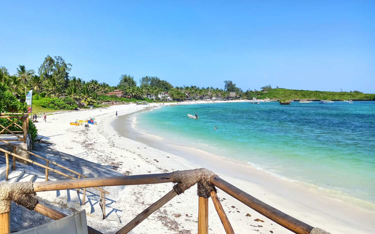View of Watamu Beach in Kenya showing a curved white sandy shoreline lined with palm trees, clear turquoise waters, small boats, and a bright blue sky, as seen from a wooden deck.