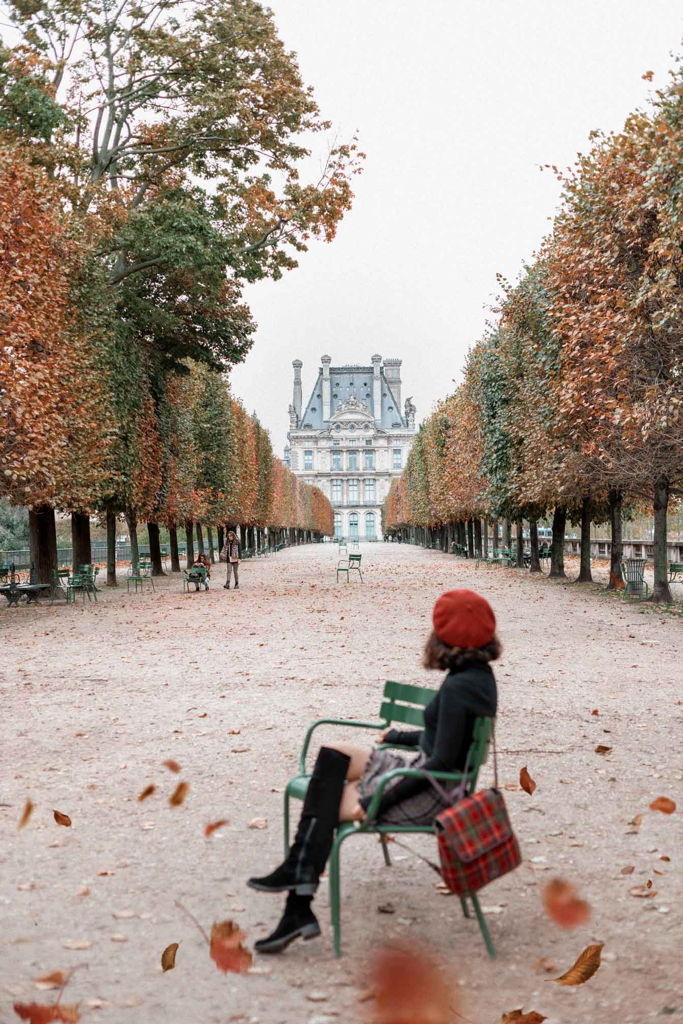 Woman in fall outfit sitting on a green bench in the Jardin des Tuileries, Paris, with autumn leaves and tree-lined path leading to the Louvre Palace in the distance.