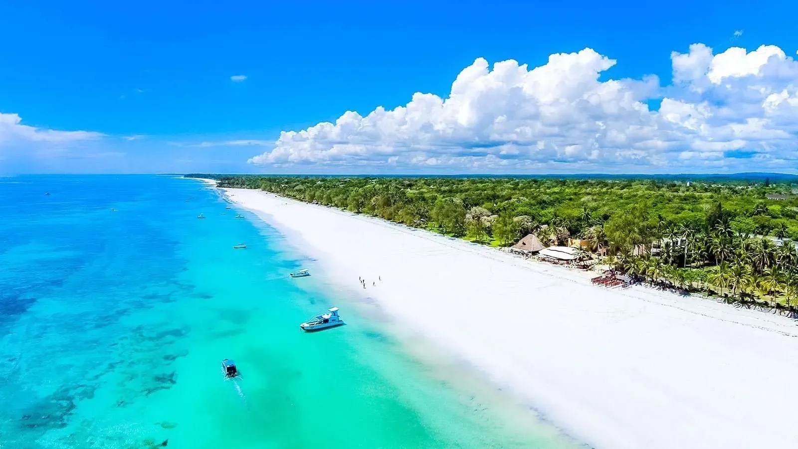 Aerial view of Diani Beach, Kenya, featuring a long stretch of white sand, lush coastal vegetation, and vibrant turquoise ocean waters under a vivid blue sky with scattered clouds.