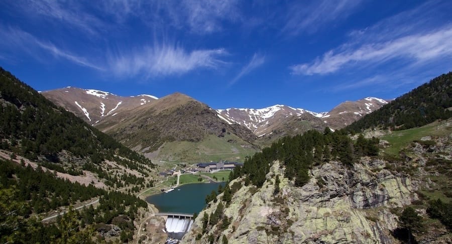 Mountain landscape in the Pyrenees with a dam, alpine lake, and snow-capped peaks, surrounded by green forests under a clear blue sky in early fall.