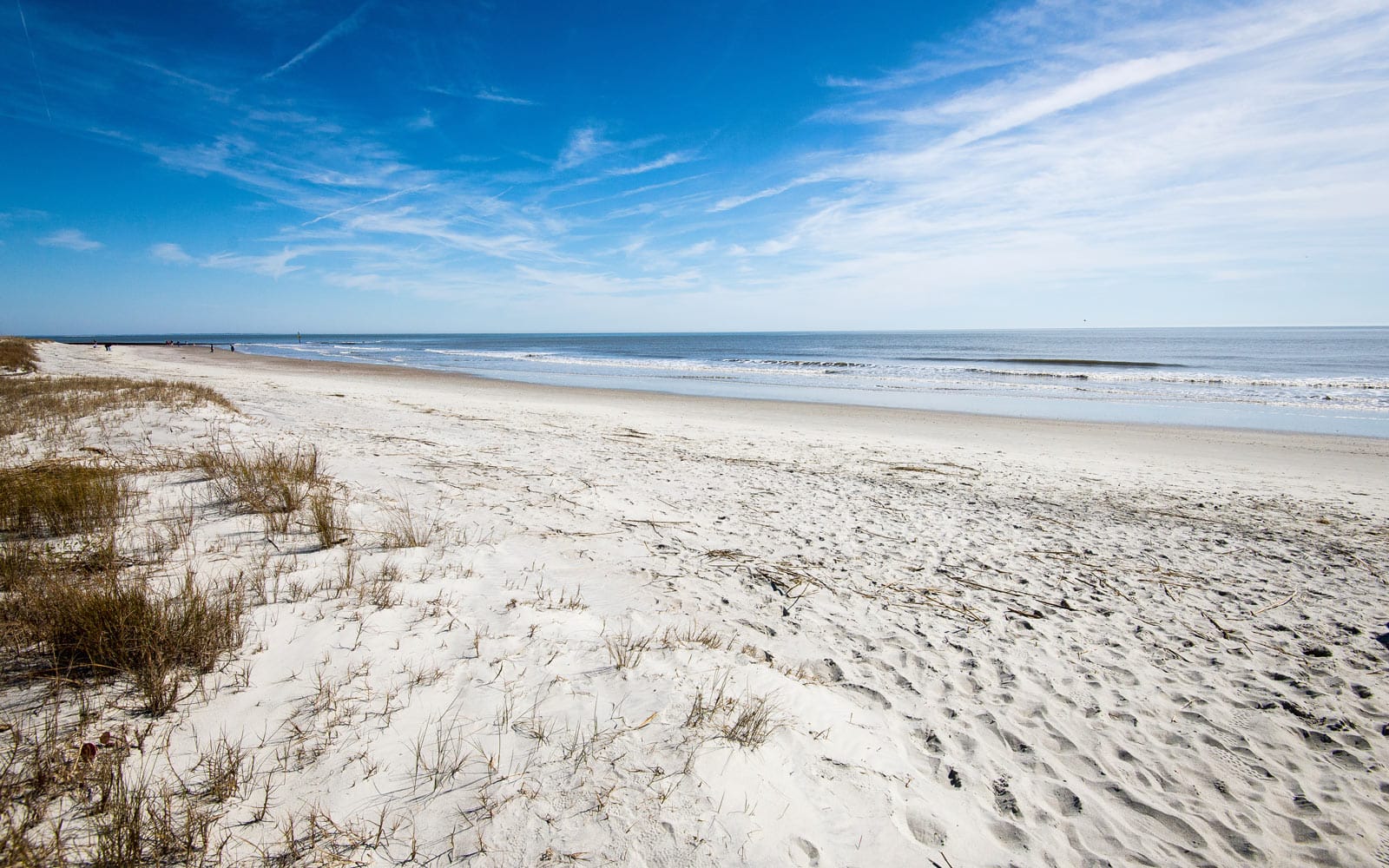 Scenic view of Hunting Island Beach in South Carolina, showing a pristine stretch of white sand, sparse dune grass, and gentle ocean waves under a clear blue sky.