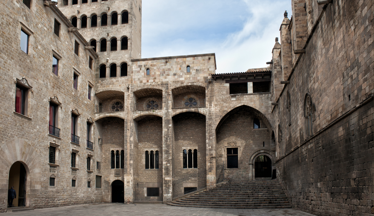 A medieval square surrounded by historic buildings, including the Palau Reial Major and the City History Museum.