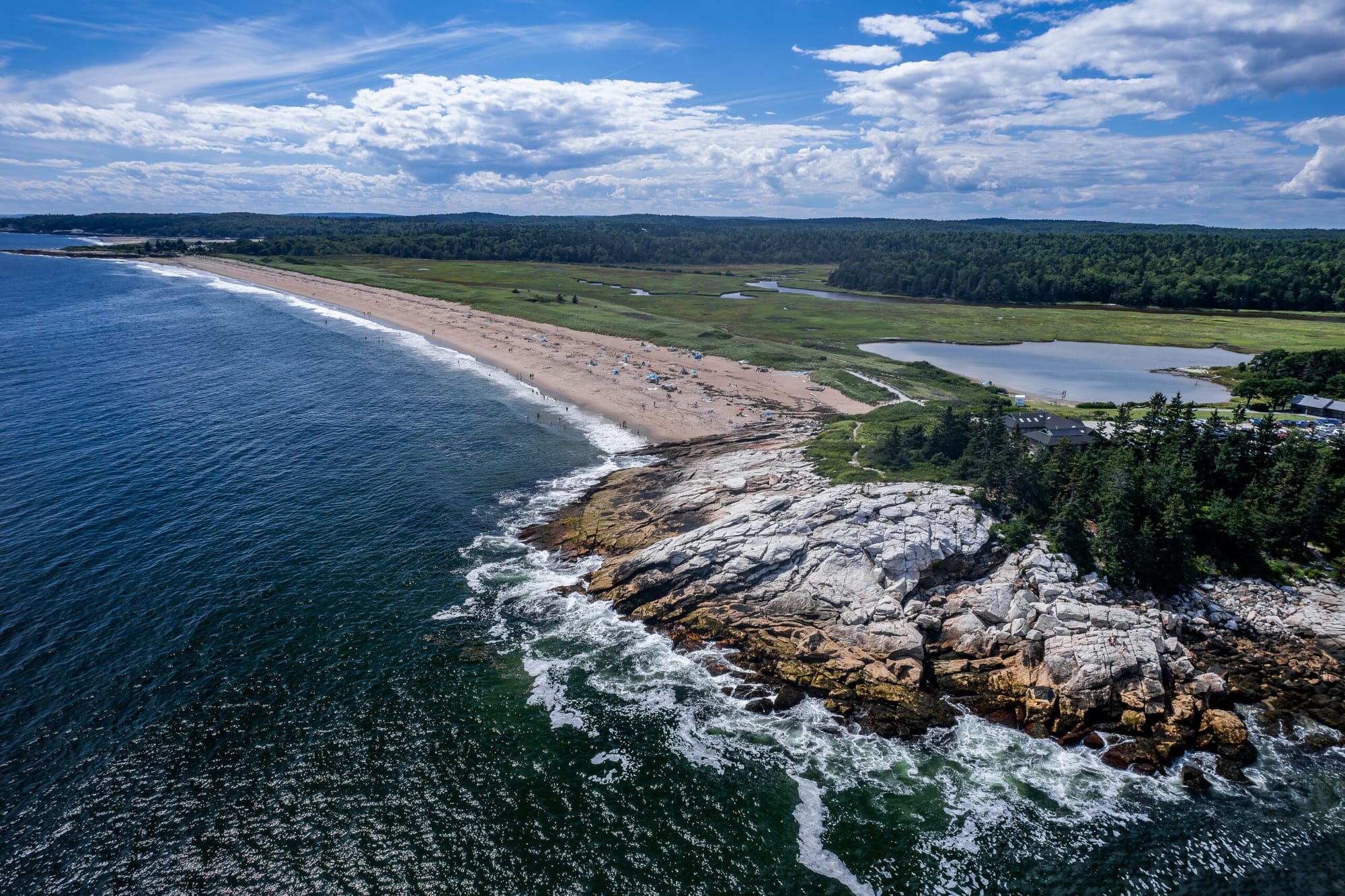  Dramatic aerial view of Reid State Park in Maine showcasing a stretch of beach bordered by dark green forest, tidal pools, rocky cliffs, and the vast open Atlantic Ocean.