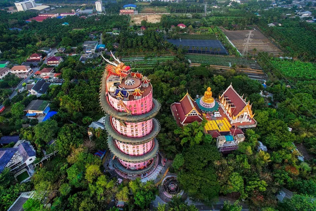Aerial view of Wat Samphran, a tall pink cylindrical temple with a giant green dragon sculpture spiraling around it, surrounded by lush greenery and traditional Thai architecture in Nakhon Pathom, Thailand.