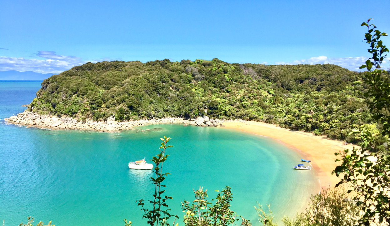Golden sand beach and turquoise bay framed by forested hills at Abel Tasman National Park in New Zealand, with anchored boats in the water.