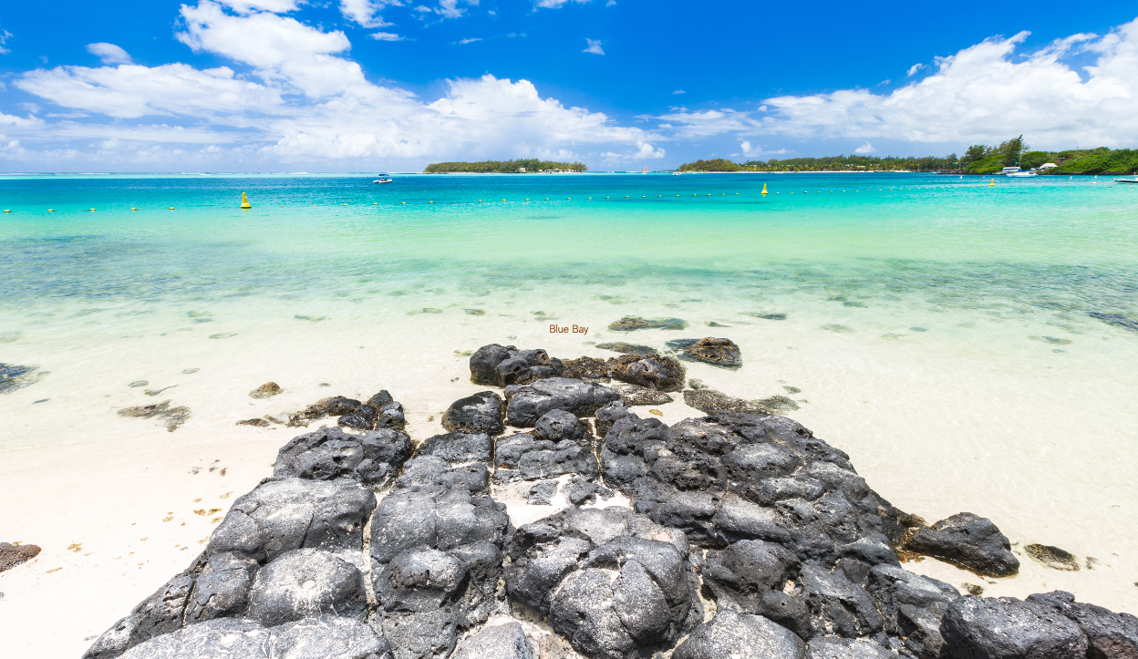 View of Blue Bay Beach in Mauritius with black volcanic rocks on white sand and clear turquoise water under a bright blue sky with scattered clouds.